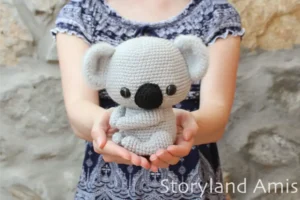 A girl holds a crocheted amigurumi koala in her hands, held out in front of her body. The girl stands in front of a rock wall and is wearing a blue and white shirt.