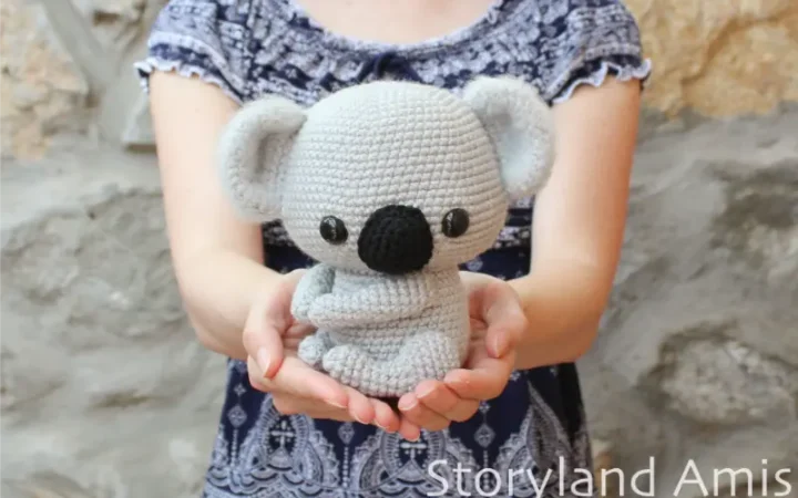 A girl holds a crocheted amigurumi koala in her hands, held out in front of her body. The girl stands in front of a rock wall and is wearing a blue and white shirt.