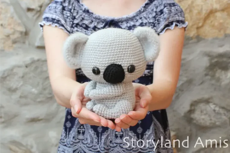 A girl holds a crocheted amigurumi koala in her hands, held out in front of her body. The girl stands in front of a rock wall and is wearing a blue and white shirt.