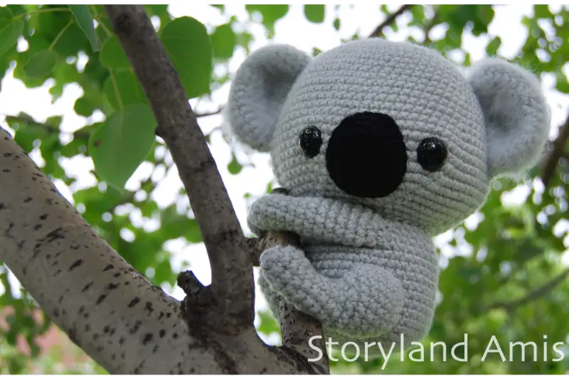 A crocheted amigurumi koala hangs on a tree branch outside with green leaves behind it.