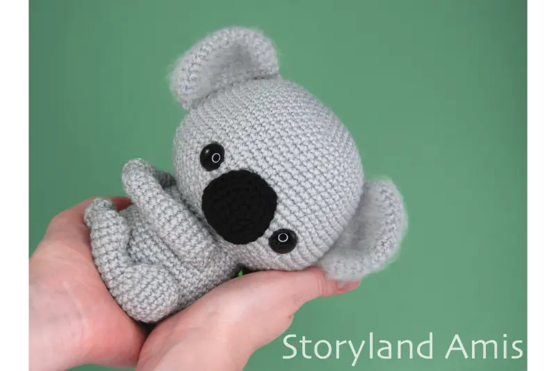 Two hands hold a crocheted amigurumi koala in front of a green background.