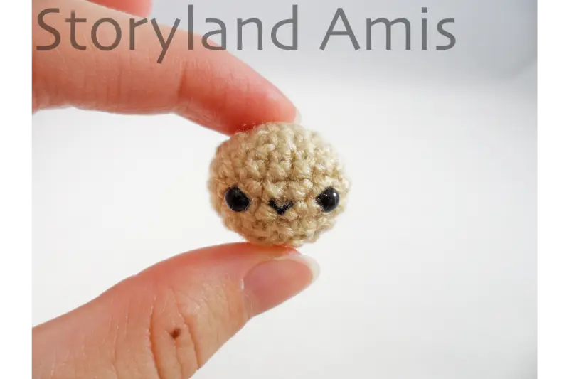 A hand holds up a tiny, crocheted doll head with an embroidered mouth and plastic safety eyes.