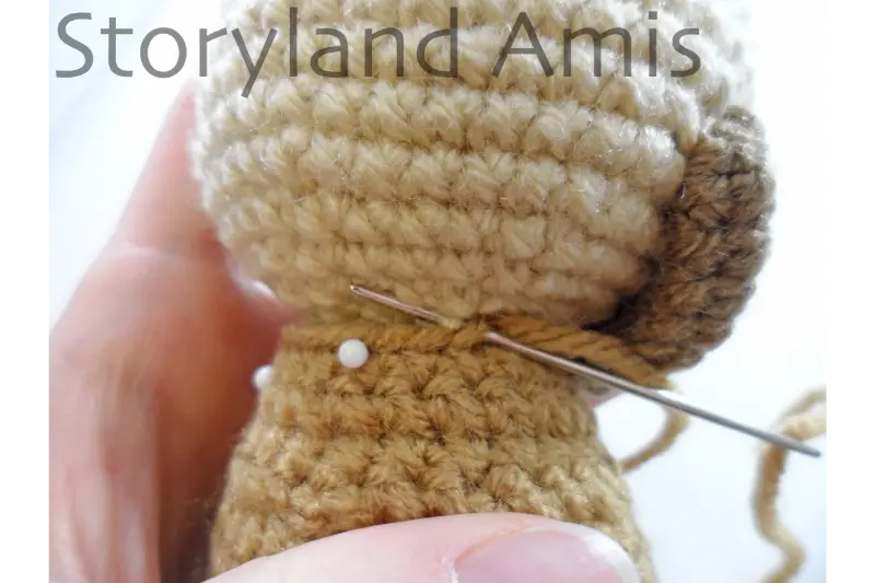 A close-up of a crocheted coat being sewn to the body of an amigurumi doll using a tapestry needle.