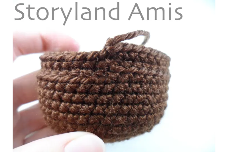 A hand holds up a brown crocheted headdress for an amigurumi doll.
