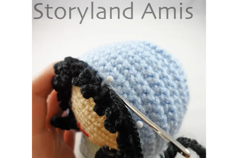 A close-up of a headdress being sewn to the head of an amigurumi doll using a tapestry needle.