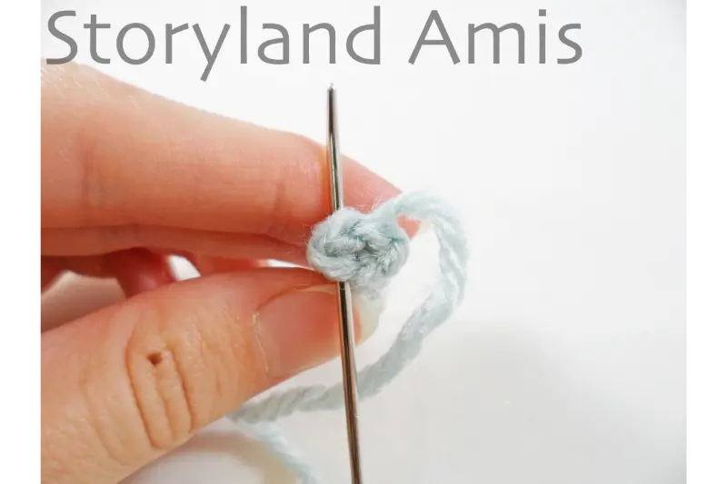 A hand holds a small crocheted doll arm. A tapestry needle is inserted in the top of the arm.