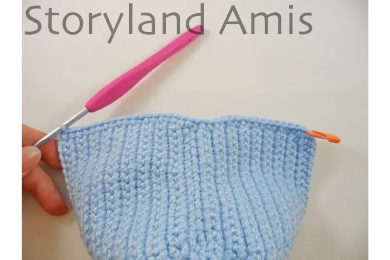 A pink crochet hook is inserted into a corner stitch of a blue crocheted headdress for an amigurumi Mary doll.