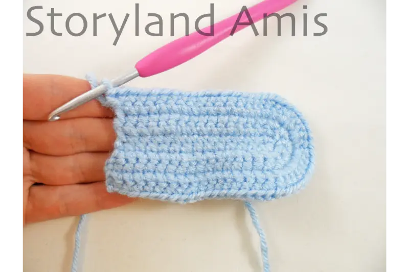 A hand holds a partially crocheted headdress for an amigurumi doll. A pink crochet hook is inserted in the corner stitch of the piece.