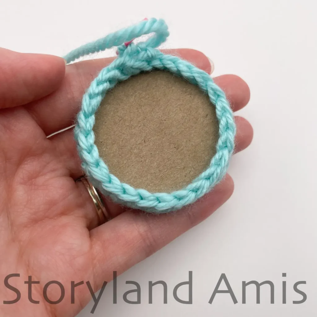 A hand holds a teal crocheted circle with a piece of cardboard inside it.