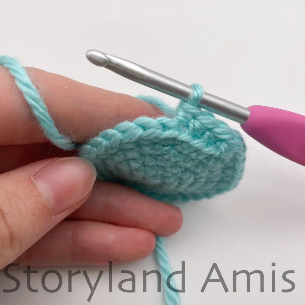 A hand holds a teal crocheted circle steady. The crocheted circle has a pink crochet hook inserted in one of the stitches.