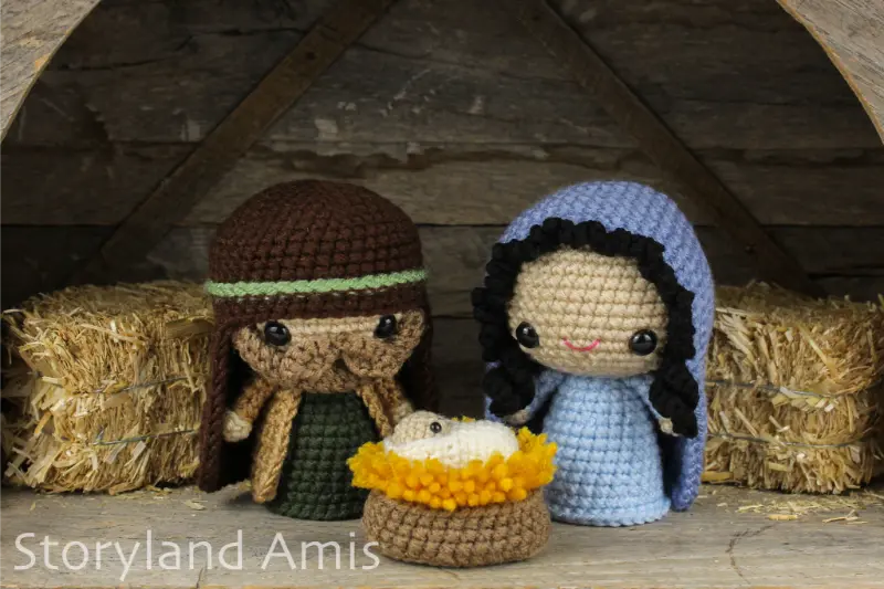 A crocheted amigurumi nativity set including Joseph, Mary, and baby Jesus is displayed in a wooden stable with small hay bales.