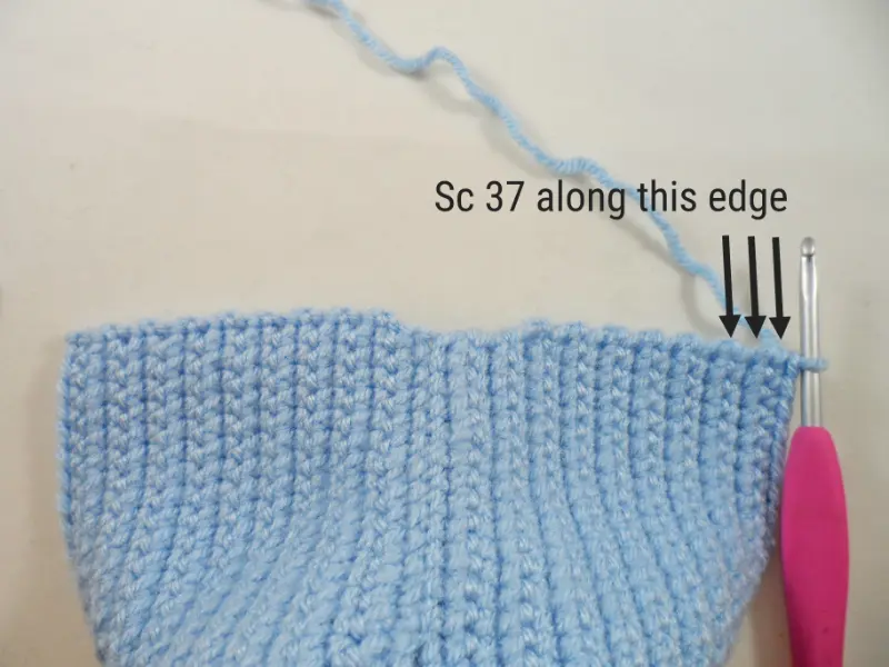A close-up of the bottom edge of a crocheted headdress for an amigurumi doll. A pink crochet hook is inserted in the corner stitch of the headdress. Text on the picture reads "sc 37 along this edge".