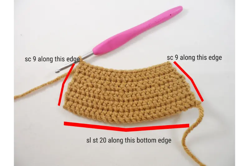 A brown crocheted rectangular piece lays on a white table with a pink crochet hook inserted in one corner.