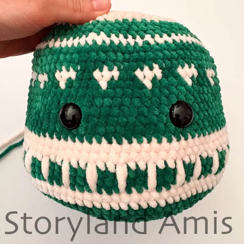 A partially crocheted amigurumi ornament is shown from the front. Two plastic safety eyes are placed on the front of the ornament.