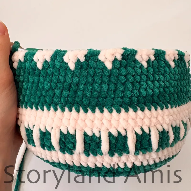 A hand holds a large, partially crocheted green amigurumi ornament with a white design.