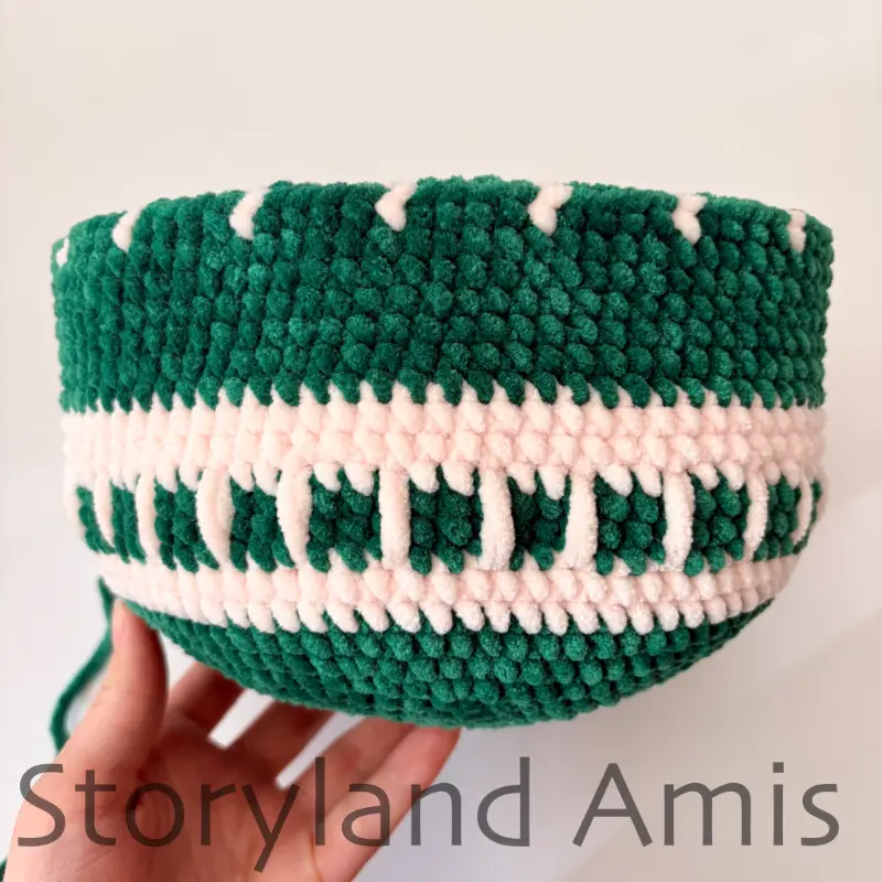 A hand holds up a large, partially crocheted green amigurumi ornament with a white design.