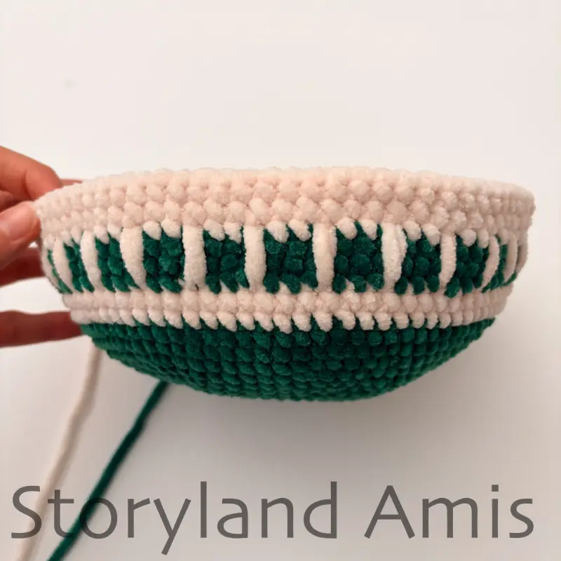 A partially crocheted round amigurumi ornament is shown against a white background while a hand holds it steady.