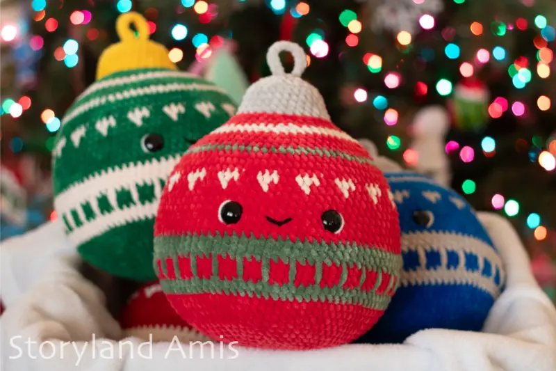 Three large amigurumi Christmas ornaments sit in a blanket-lined basket. A Christmas tree is in the background. 