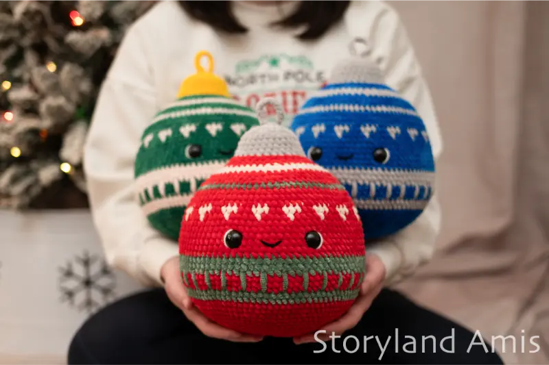 A girl sits in front of a Christmas tree holding three large, plush crocheted Christmas ornaments.