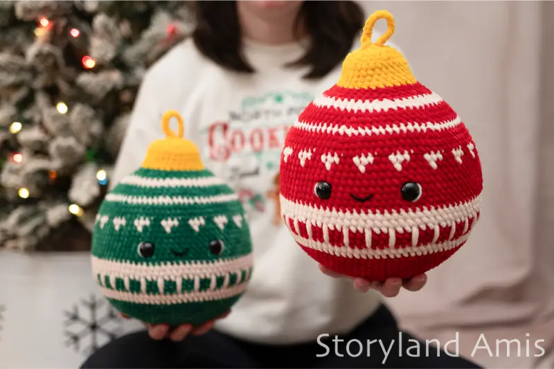 A girl sits in front of a Christmas tree holding two large, plush crocheted Christmas ornaments.