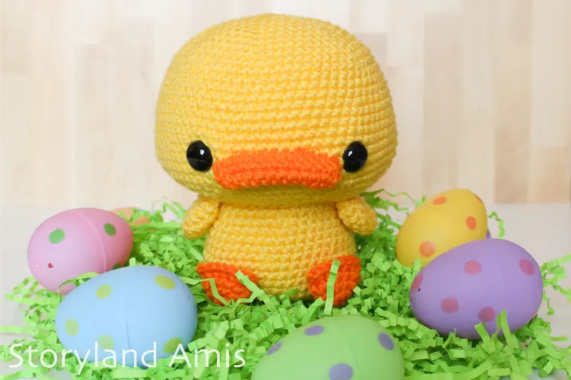 A crocheted amigurumi duck sits on green paper Easter grass with colorful plastic Easter eggs around it.