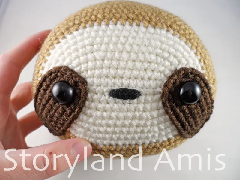 A hand holds a crocheted amigurumi sloth head.