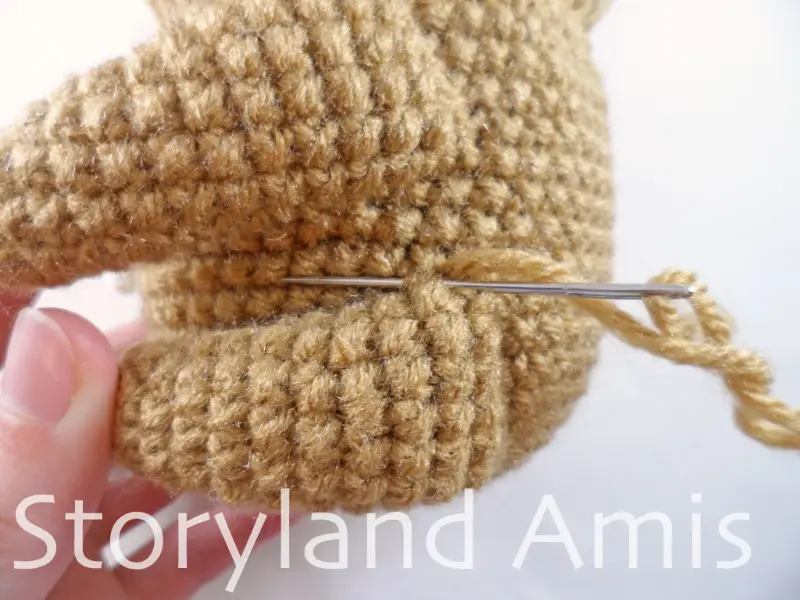 A close-up of a tapestry needle being used to sew a crocheted leg to the body of an amigurumi sloth.