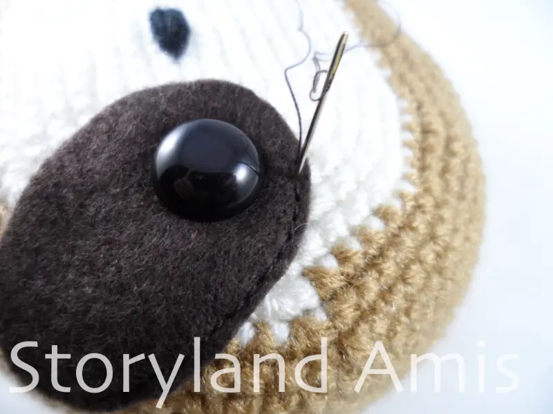 A close-up of a needle and thread being used to sew a brown felt eye patch to an amigurumi sloth's head.