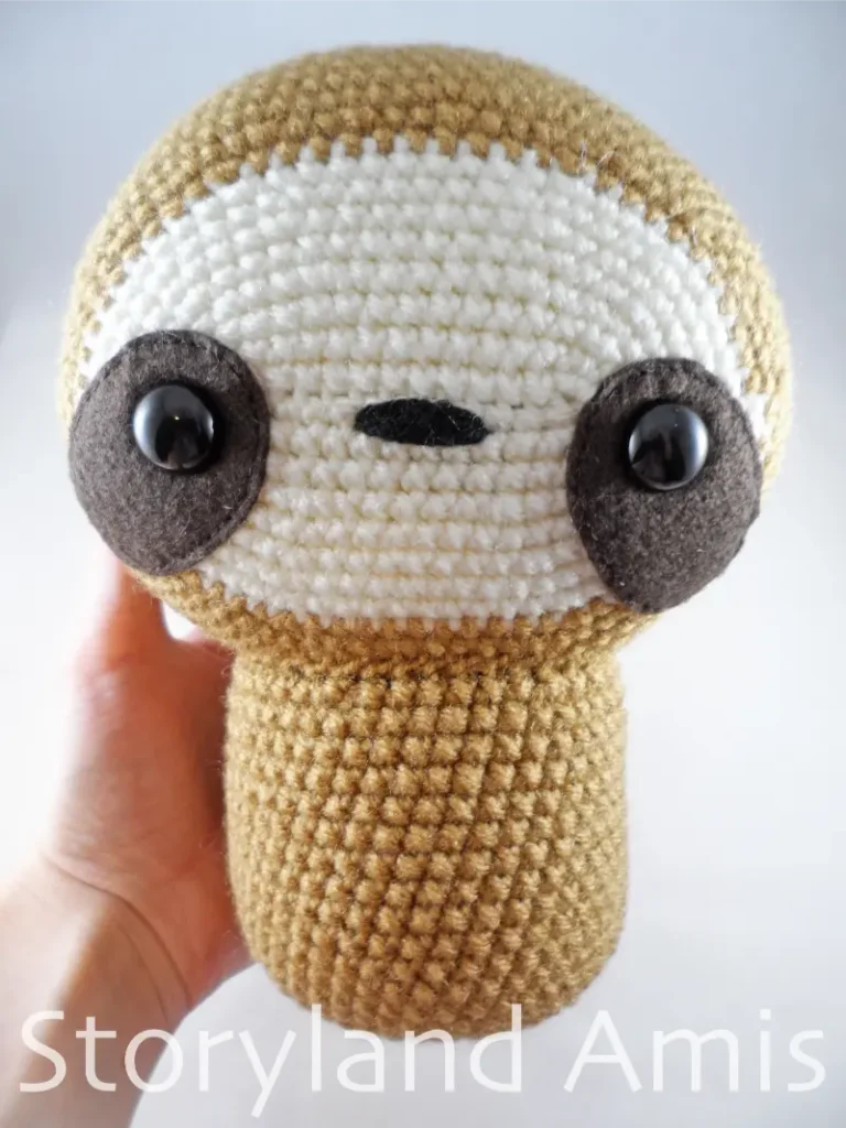 A hand holds a partially finished crocheted amigurumi sloth.