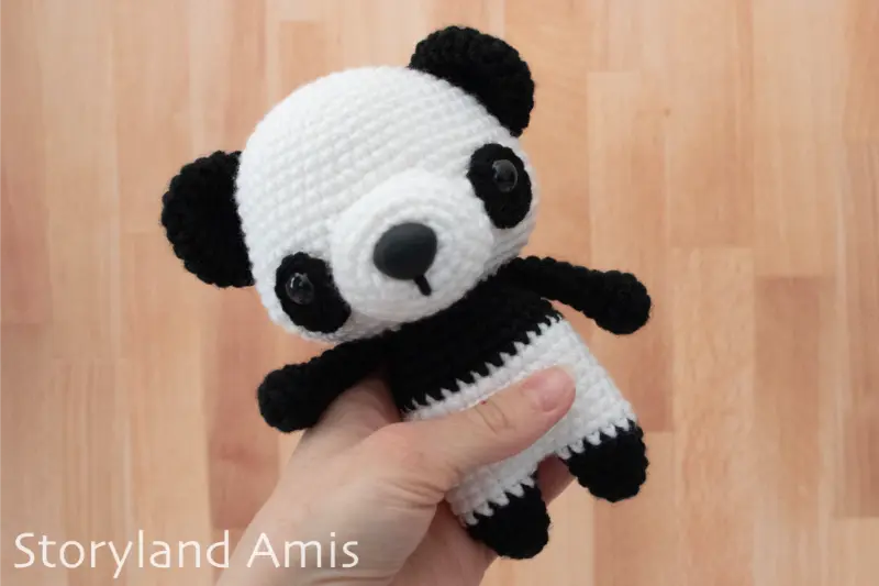 A hand holds a crocheted black and white panda amigurumi toy against a wood background.