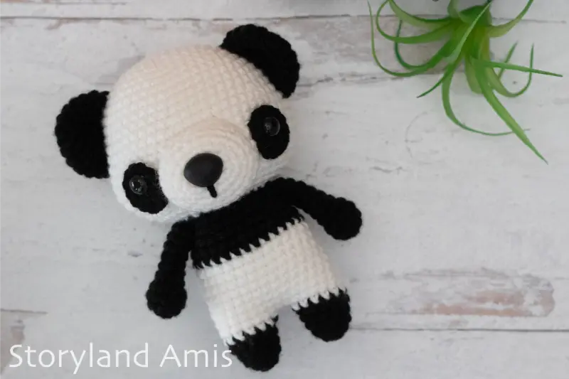 A crocheted black and white amigurumi panda toy lies on a light wood table. A green plastic plant is in the upper right corner of the picture.