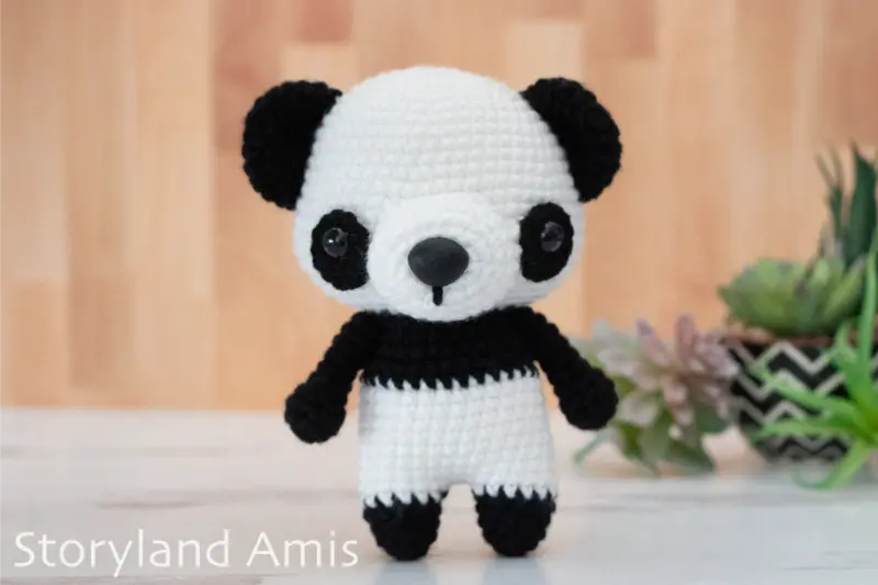 A crocheted black and white amigurumi panda stands on a light wood table with fake green plants in the background.