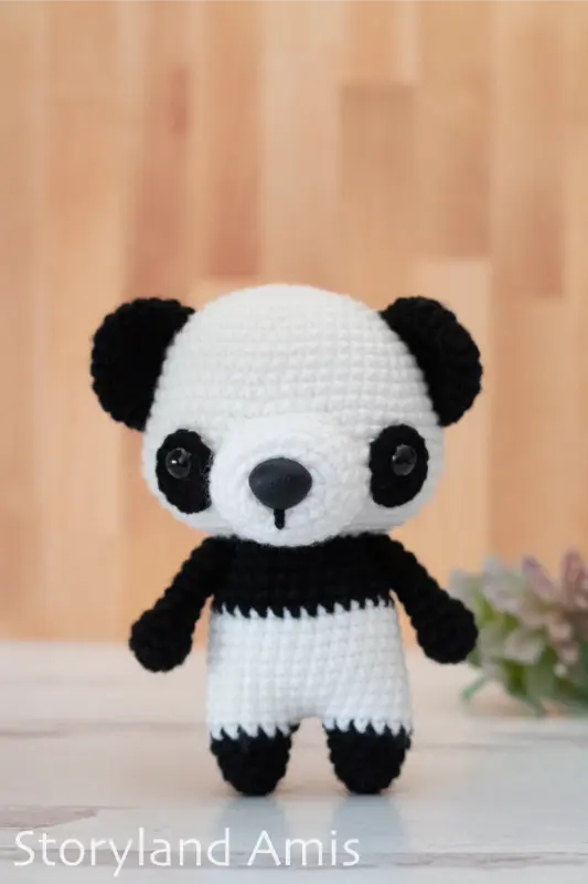 A crocheted black and white amigurumi panda stands on a light wood table with fake green plants in the background.