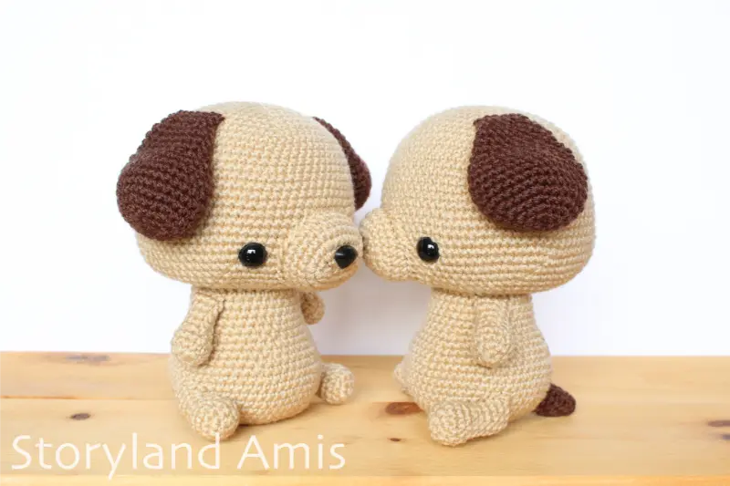 Two tan and brown amigurumi puppy dogs sit, side by side, on a wooden table.