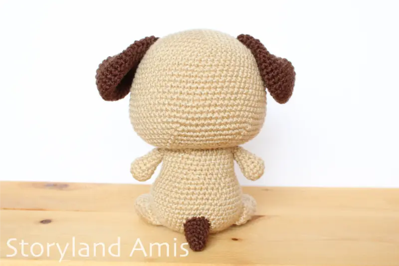 A tan and dark brown amigurumi puppy dog sits on a wooden table.