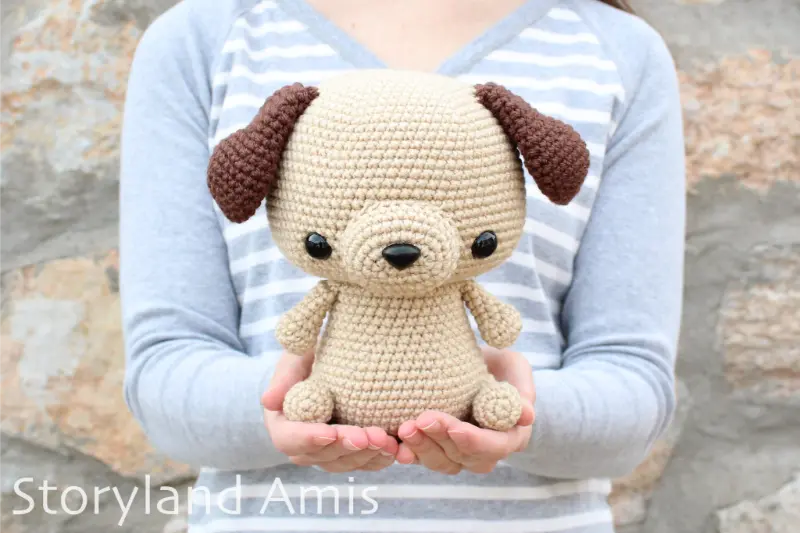 A girl stands in front of a brick wall wearing a gray and white striped shirt holding a brown crocheted amigurumi puppy dog. 