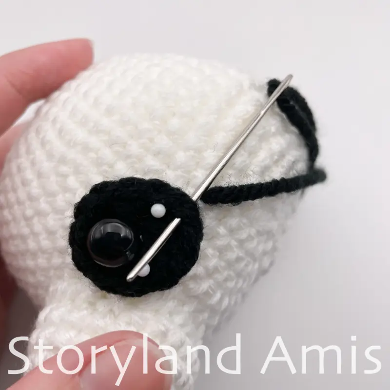 A close-up showing a hand using a silver tapestry needle to sew a crocheted black eye patch to an amigurumi panda's head.