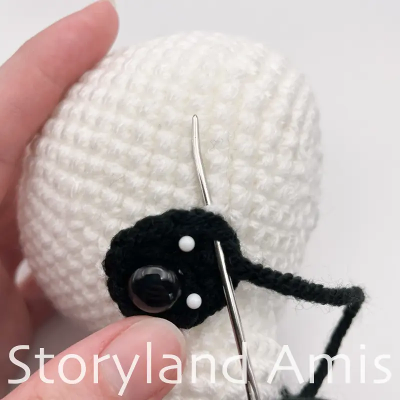 A close-up showing a hand using a silver tapestry needle to sew a crocheted black eye patch to an amigurumi panda's head.