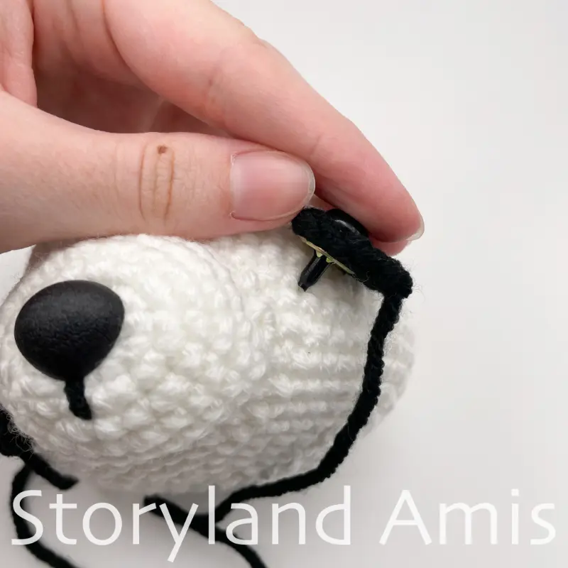 A close-up of a hand wiggling the post of a safety eye in between the stitches on an amigurumi panda's head.