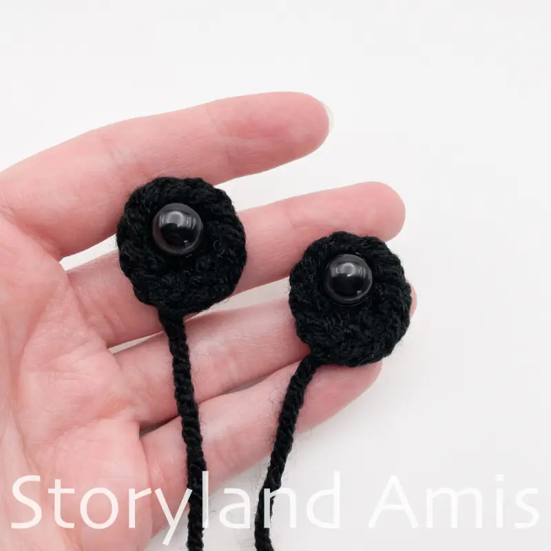 A hand holds two black crocheted eye patches with black plastic safety eyes inserted in the center of each.