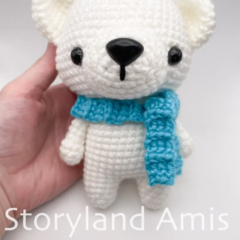 A hand holds an amigurumi polar bear wearing a blue crocheted scarf.