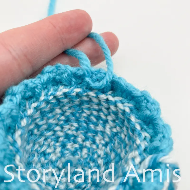 A close-up of two yarn tails knotted together on the edge of a small blue crocheted stocking cap.
