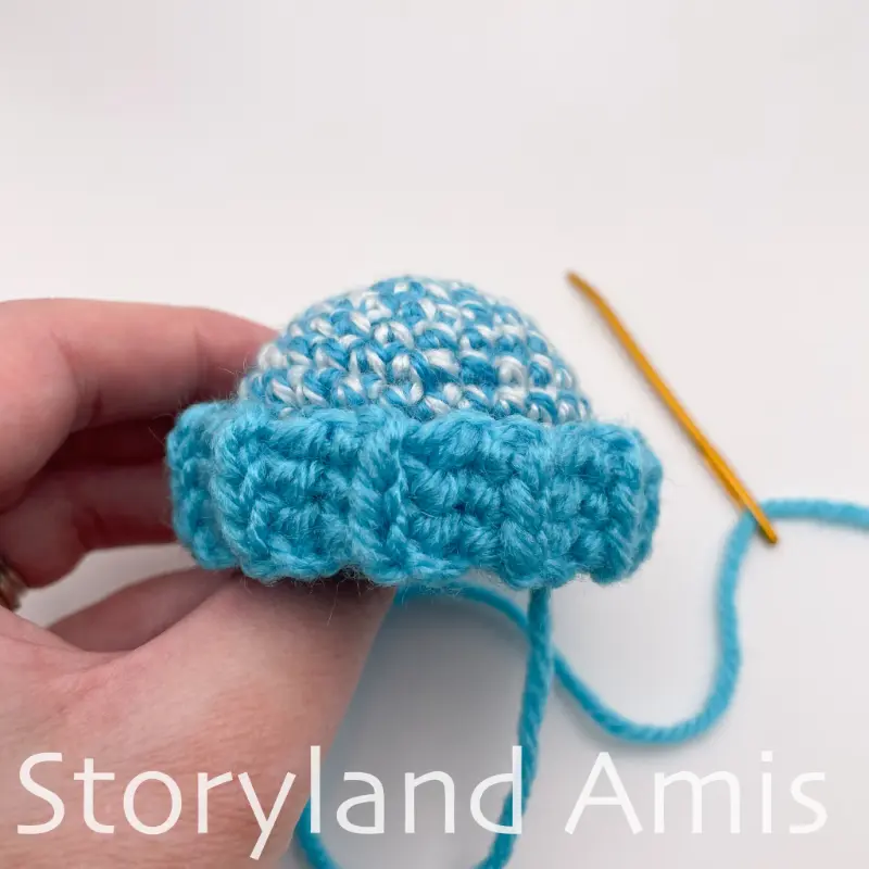 A hand holds a partially finished small blue crocheted stocking cap. 