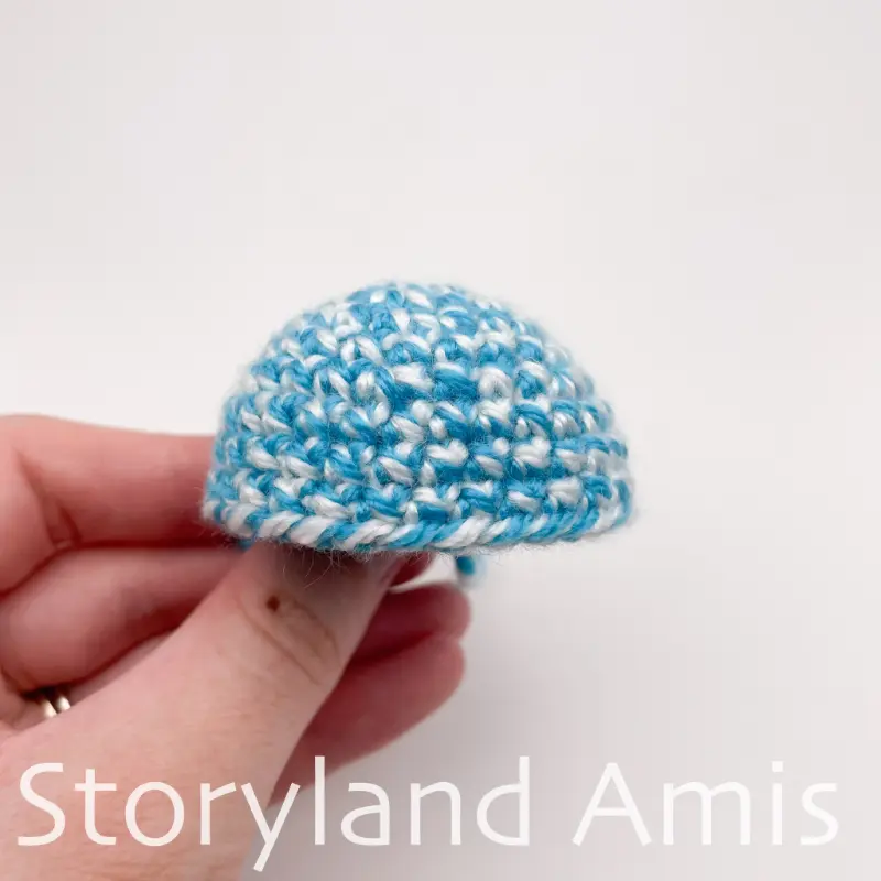 A hand holds a small, partially finished crocheted stocking cap made with blue and white variegated yarn. 