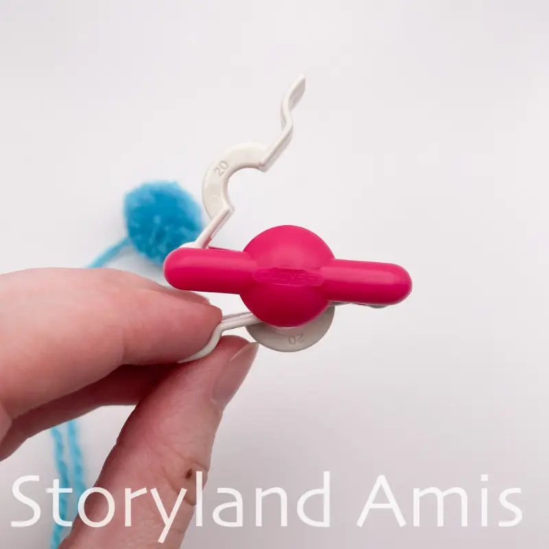 A hand holds a small plastic pink and white pom pom maker.