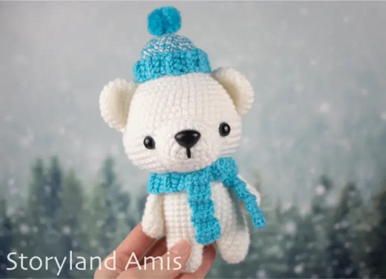 A hand holds an amigurumi polar bear wearing a blue crocheted hat and scarf.