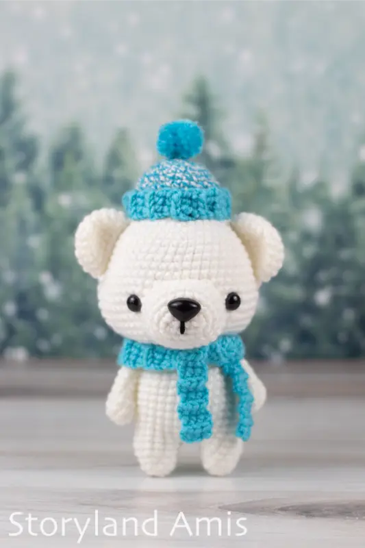 An amigurumi polar bear wearing a crocheted blue scarf and hat stand on a wooden table in front of a snowy tree background. 