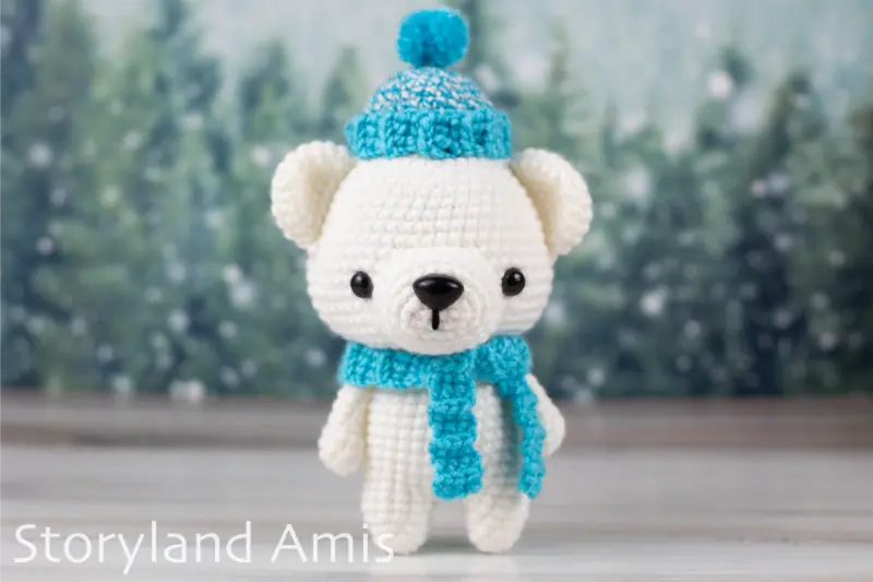 An amigurumi polar bear wearing a crocheted blue scarf and hat stand on a wooden table in front of a snowy tree background.