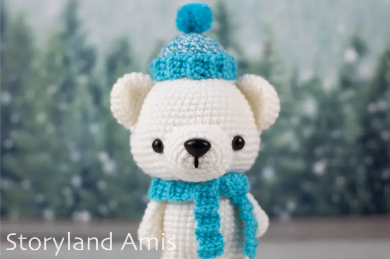 An amigurumi polar bear wearing a crocheted blue scarf and hat stand on a wooden table in front of a snowy tree background.
