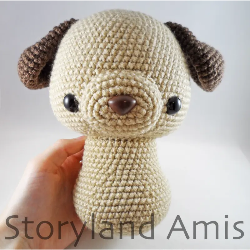 A hand holds a partially finished crocheted tan and brown amigurumi dog.